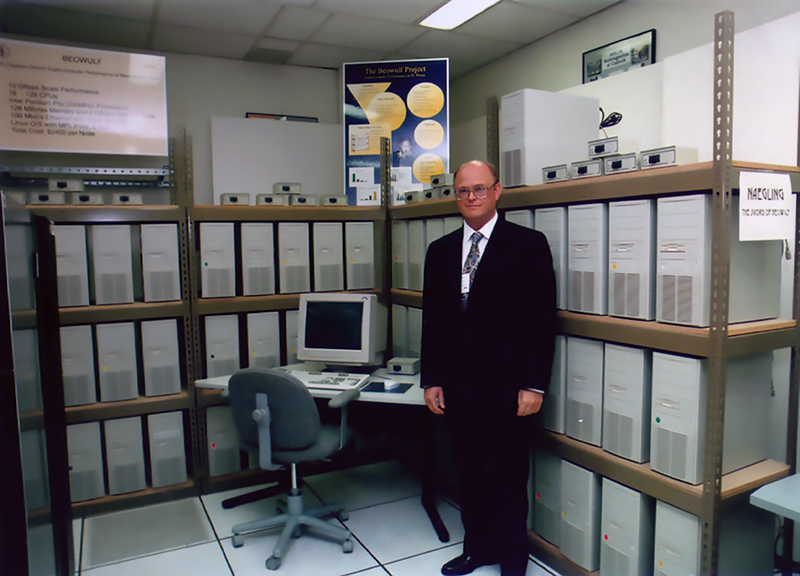 Thomas Sterling stands by a PC on a desk with dozens of CPUs on shelves behind him