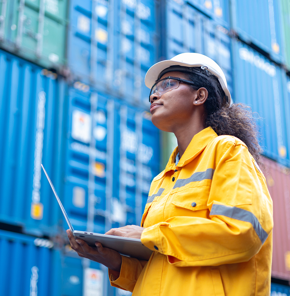 A woman holds a laptop in front of stacked shipping containers