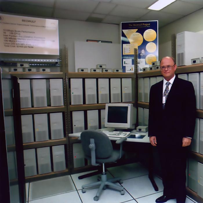 Thomas Sterling stands by a PC on a desk with dozens of CPUs on shelves behind him