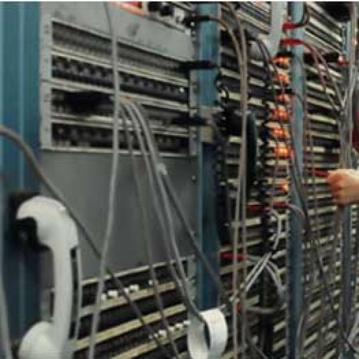 Two men plug and unplug wires attached to a wall of ports. Digital system covers the wall with no visible cables