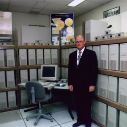 Thomas Sterling stands by a PC on a desk with dozens of CPUs on shelves behind him