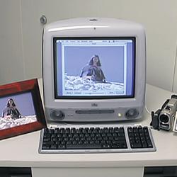 An imac with an image on the screen, a framed image and a video camera beside it.